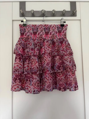 Rosa volangkjol med blommönster - Söt kortkjol i rosa toner med blommigt mönster och flera volanglager. Kjolen har en bred, smockad midja som ger en bekväm passform och ett luftigt fall. Perfekt för dig som gillar färg och mönster!