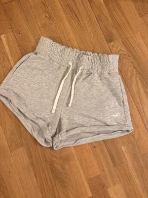 Grå mjukisshorts från Hollister XS - Säljer ett par ljusgrå mjukisshorts från Hollister i storlek XS. Shortsen har hög midja med bred resår och vita dragsnören samt sidofickor. Tillverkade i mjukt sweatshirtmaterial, perfekta för chill dagar.