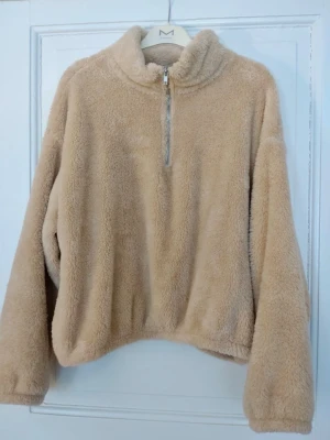 Beige fluffig halvzip tröja - Mysig beige stickad tröja med fluffig teddystruktur och halv dragkedja framtill. Tröjan har hög krage och långa ärmar, perfekt för kalla dagar. Passar dig som gillar en avslappnad och trendig stil.