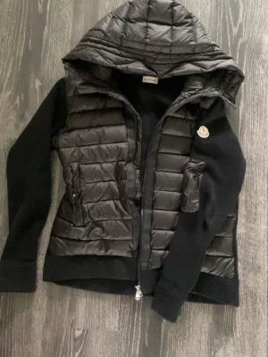 Svart pufferjacka från Moncler - Snygg svart pufferjacka från Moncler med quiltad front och huva. Jackan har stickade ärmar och sidor, dragkedja framtill och två fickor med dragkedja. Ikonisk Moncler-logga på ärmen. Perfekt för kalla vinterdagar.