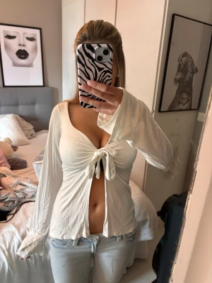 Design By Si knyt tröja - Trendig vit långärmad topp med djup v-ringning och knyt framtill. Toppen är croppad och har vida ärmar som ger en avslappnad vibe. Perfekt att styla med jeans för en cool och enkel look.