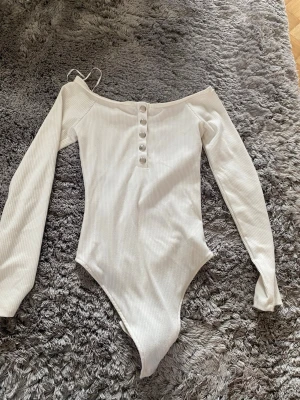 Vit ribbad off shoulder body - Snygg vit ribbad body med off shoulder-design och långa ärmar. Framtill finns dekorativa knappar som ger en extra detalj. Perfekt att styla med jeans eller kjol för en trendig look.