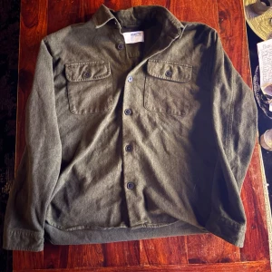 Olivgrön overshirt med bröstfickor - Stilren olivgrön overshirt i mjukt tyg med klassisk krage och knäppning framtill. Två bröstfickor med lock och knappar ger en schysst utility-vibe. Perfekt lager på lager-plagg för dig som gillar avslappnad men snygg stil.
