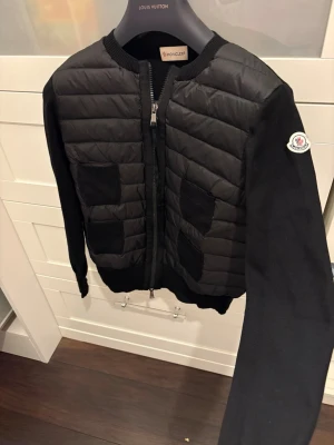 Svart cardiganjacka från Moncler - Snygg svart cardiganjacka från Moncler med quiltad front och stickade ärmar. Jackan har fyra ribbade fickor framtill och dragkedja. Moncler-logga på ena ärmen. Perfekt för lager-på-lager och stilren look. Köpt på vinted med äkthetsverifikationen men knappt använd så i nyskick!