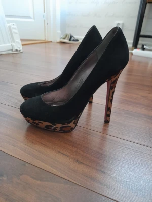 Svarta pumps med leopardklack - Snygga svarta pumps från Paris Hilton med hög platå och klack i leopardmönster. Skorna har rundad tå och är tillverkade i mockaliknande material. Perfekta för dig som vill sticka ut med en cool detalj på klacken.