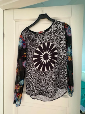 Mönstrad långärmad topp Desigual - Unik långärmad topp från Desigual med svartvitt grafiskt mönster och färgglada blommiga ärmar. Rund halsringning och asymmetrisk nederkant ger en cool look. Perfekt för dig som gillar statement-plagg och vill sticka ut.