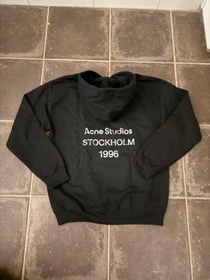 Svart hoodie Acne Studios Stockholm - Svart hoodie från Acne Studios med trycket 'Acne Studios STOCKHOLM 1996' i vitt på ryggen. Klassisk passform med huva och ribbade muddar. OBS! Osäker på äktheten därav priset!! Pris går att diskutera vid snabb affär!!