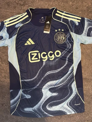 Ajax blå matchtröja Adidas - Säljer en blå Ajax matchtröja från Adidas med ljusblå och gula detaljer. Tröjan har Ajax klubbmärke, tre stjärnor och Ziggo-logga på bröstet. Tillverkad i ett lätt och ventilerande material med Aeroready-teknologi. Perfekt för fotbollsträning eller match.
