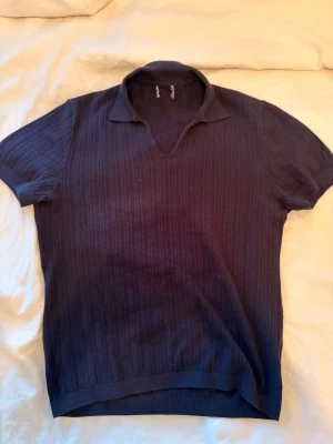 Mörkblå ribbad polotopp - Snygg mörkblå ribbad polotopp med korta ärmar och öppen krage. Toppen har en klassisk, lite loose passform och är tillverkad i ett mjukt stickat material som känns skönt mot huden. Perfekt för dig som gillar stilrena och tidlösa plagg.