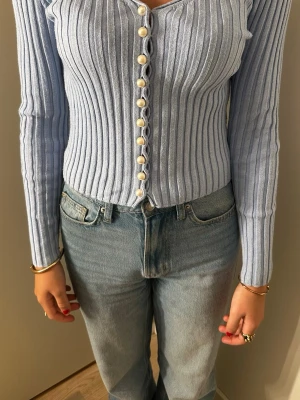 Ljusblå ribbad stickad cardigan - Säljer en ljusblå ribbad stickad cardigan med långa ärmar och v-ringning. Tröjan har dekorativa pärlknappar framtill och är figurnära i modellen. Perfekt att styla med jeans för en trendig look.