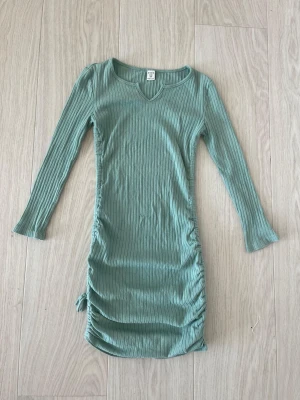 Grön ribbad klänning - Snygg grön ribbad klänning med lång ärm och v-ringning. Klänningen har en tight passform och snörning i sidan som ger en cool detalj. Perfekt för dig som gillar trendiga och bekväma plagg.