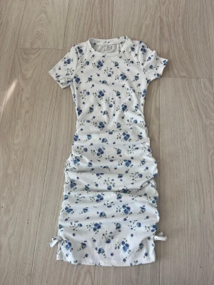 Vit blommig klänning med knyt - Supersöt vit sommarklänning med blå blommor och ribbad struktur. Klänningen har korta ärmar, rund halsringning och knytband i sidorna nedtill för en snygg detalj. Perfekt för sommarens alla tillfällen!