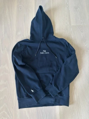 Mörkblå hoodie från Polo Ralph Lauren - Snygg mörkblå hoodie från Polo Ralph Lauren med broderad logga på bröstet och liten vit häst på ärmen. Klassisk känguruficka framtill och justerbar huva med dragsko. Perfekt för en chill och stilren look. Vid intresse är det bara att höra av sig. 
