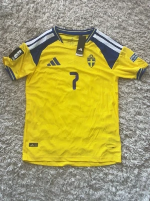 Svensk landslagströja nr 7  - Gul svensk landslagströja med blå detaljer, nummer 7 och namnet 'Bergvall' på ryggen. Tröjan har broderad landslagssköld, tryckta loggor och är tillverkad i ett lätt, ventilerande funktionsmaterial. Perfekt för fotboll eller att bära på matchdag.