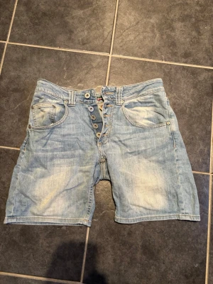 Ljusblå jeansshorts med knappar - Säljer ett par ljusblå jeansshorts med flera knappar framtill och klassiska fem fickor. Shortsen har en avslappnad passform och är perfekta för varma dagar. Snygg slitning och detaljerad söm på bakfickorna.