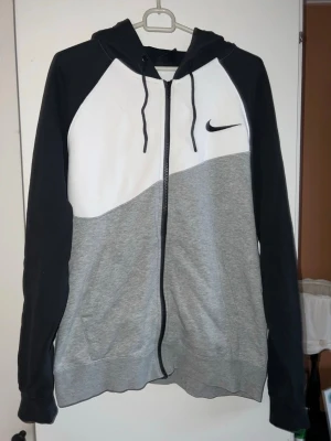 Nike tröja - Nike hoodie med dragkedja i svart, grå och vit färgblock. Stor Nike-logga på ryggen och mindre logga på bröstet. Huva med dragsko och två fickor framtill. Tillverkad i mjukt sweatshirtmaterial, perfekt för en avslappnad stil.