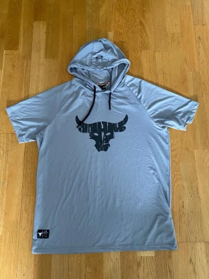 Ljusgrå hoodie med tjurtryck - Snygg ljusgrå hoodie med korta ärmar och svart tjurtryck på bröstet. Huvan har svarta dragsnören och materialet känns lätt och sportigt, perfekt för en avslappnad stil. Passar dig som gillar streetwear och vill sticka ut lite extra.