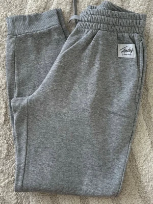 Grå mjukisbyxor från Andy by Frank Dandy - Snygga grå mjukisbyxor från Andy by Frank Dandy med resår i midjan och muddar vid bensluten. Tillverkade i mjukt och bekvämt material, perfekta för chill dagar. Klassisk passform och diskret logga på ena benet.