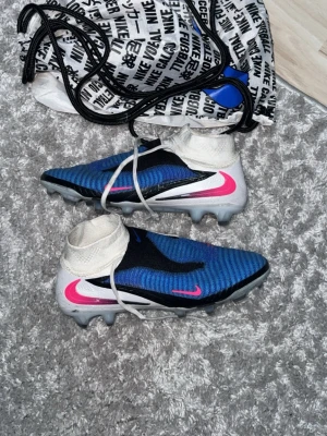 Nike phantom elite - Strl 41! Använda ca 4 gånger. Tvättar bort det som är smutsigt innan köp!!Säljer ett par Nike fotbollsskor i blått med vita och svarta detaljer samt rosa swoosh. Skorna har inbyggd strumpa för extra stöd runt ankeln och dobbar för spel på gräs. Materialet är syntetiskt och designen är modern och sportig. Perfekt för dig som vill sticka ut på planen.