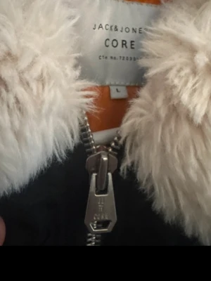 Jack & Jones svart bomberjacka L - Snygg svart bomberjacka från Jack & Jones med fluffig vit fuskpäls runt kragen och orange innerfoder. Jackan har en cool detalj med röd 'Remove Before Flight'-tag på ärmen och grov dragkedja framtill. Perfekt för dig som vill sticka ut.