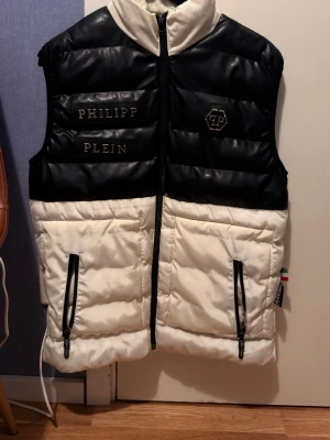 Svartvit dunväst från Philipp Plein - Snygg dunväst i svart och vitt från Philipp Plein. Västen har hög krage, två dragkedjefickor fram och coola detaljer med logga och broderad text. Materialet är glansigt och quiltat, perfekt för lager på lager och streetwear-vibe.