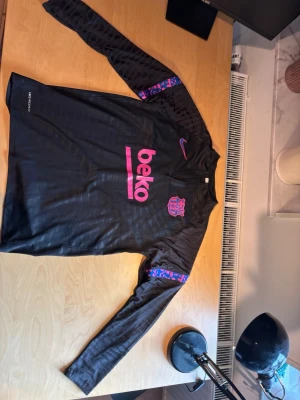 Barca långärmad  - Snygg svart långärmad träningströja från Nike med FC Barcelona-logga och rosa detaljer. Tröjan har trycket 'beko' på bröstet och mönstrade band i lila och rosa på ärmarna. Perfekt för fotbollsträning eller som supporterplagg.