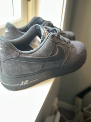 Mörkgrå Nike Air Force 1 sneakers - Säljer ett par mörkgrå Nike Air Force 1 sneakers i mocka med klassisk swoosh på sidan och AIR-detalj på sulan. Skorna har lågt skaft och matchar lätt till olika outfits. Perfekta för dig som gillar stilrena och tidlösa sneakers. Köpta i London på JD-sports under semester. Var inte riktigt min smak insåg jag sen. Använda 5-10 gånger. Kom med prisförslag😀