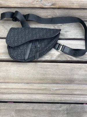 Svart axelväska i sadelmodell - Snygg svart axelväska i sadelmodell med präglat mönster och justerbar axelrem. Väskan har en unik form och detaljerade sömmar, samt ett diskret spänne framtill. Materialet ser ut att vara canvas med läderdetaljer.