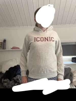 Grå hoodie med texten ICONIC - Snygg grå hoodie med huva och dragsko. På bröstet står det 'ICONIC' i stora vinröda bokstäver. Hoodien har ribbade muddar vid ärmslut och nederkant. Perfekt för en chill och avslappnad stil. Materialet känns mjukt och bekvämt, troligen bomullsblandning.