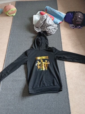 Svart Fortnite hoodie med tryck - Snygg svart hoodie med stort Fortnite-tryck på bröstet i gult och färg. Klassisk huva och långa ärmar. Perfekt för dig som gillar gaming och vill ha en bekväm och stilren tröja. Materialet känns mjukt och passar bra till vardags.