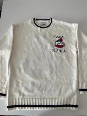 Vit stickad tröja från Casablanca - Snygg vit stickad tröja från Casablanca med broderad logga och motiv på bröstet. Tröjan har svarta ränder vid hals, ärmslut och nederkant samt ribbstickade muddar. Perfekt för dig som gillar stilrena och trendiga plagg. S men liten i storleken!