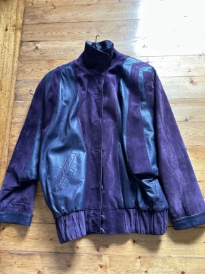 Lila retro skinnjacka - Otroligt fin vintage skinnjacka i äkta lammskinn och mocka, tillverkad i Spanien. Den djupa lila färgen tillsammans med de svarta skinnpanelerna gör den väldigt speciell – jag har själv aldrig sett en likadan. Den har den perfekta 80/90-talspassformen med markerade axlar, hög krage och resår i midjan. Känns exklusiv och har en riktigt snygg vintagekänsla. Fint skick med endast normalt slitage. Storlek 40-42 (passar även oversize). Ett unikt plagg som är svårt att hitta igen när det väl är sålt.