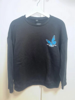 Svart sweatshirt med blå fjärilstryck - Cool svart sweatshirt med rund hals och långa ärmar. På bröstet finns ett blått fjärilstryck och texten 'This story is not finished'. På ryggen finns en stor blå fjäril och texten 'Rules are made to be broken, this story is not finished'. Mjuk och skön bomullskänsla.