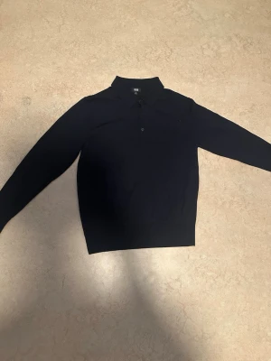 Mörkblå långärmad pikétröja - Klassisk mörkblå långärmad pikétröja med krage och knappar framtill. Tröjan har en stilren och enkel design som passar till mycket. Perfekt för dig som gillar en clean look.