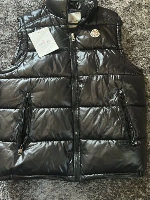 Svart dunväst från Moncler - Snygg svart dunväst från Moncler med glansig finish och klassisk logga på bröstet. Västen har två dragkedjefickor framtill och hög krage. Perfekt för lager-på-lager och ger en cool streetstyle-vibe.