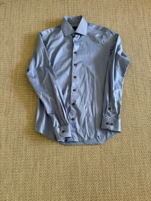 Ljusblå slim fit skjorta Dressmann - Säljer en ljusblå skjorta från Dressmann i slim fit-modell. Skjortan har långa ärmar, klassisk krage och knappar framtill samt vid manschetterna. Perfekt för dig som gillar en stilren och enkel look.
