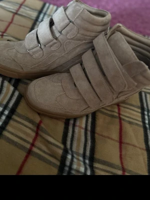 Beige sneakers med kardborreband - Snygga beige sneakers med högt skaft och flera breda kardborreband. Skorna har en chunky sula i gummi och är tillverkade i mockaliknande material. Perfekta för dig som gillar streetstyle och vill ha något unikt till outfiten.
