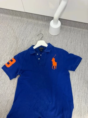 Blå pikétröja från Ralph Lauren - Klassisk blå pikétröja från Ralph Lauren med stor orange logga på bröstet och en orange siffra på ärmen. Tröjan har krage, korta ärmar och knappar vid halsen. Perfekt för en sportig och stilren look pris och byten går att diskuteras 