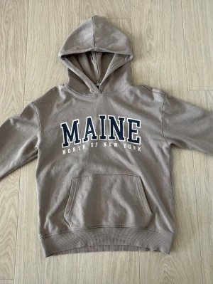 Beige hoodie med MAINE-tryck - Säljer en beige hoodie med huva och stor ficka framtill. På bröstet står det 'MAINE NORTH OF NEW YORK' i stora bokstäver. Hoodien har ribbade muddar vid ärmslut och nederkant. Perfekt för en avslappnad och trendig look.