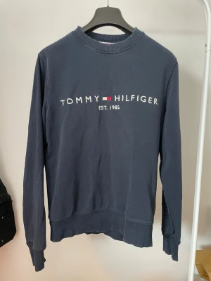 Mörkblå Tommy Hilfiger sweatshirt - Mörkblå sweatshirt från Tommy Hilfiger med klassisk logga och texten 'EST. 1985' på bröstet. Tröjan har rund halsringning, ribbade muddar och är tillverkad i mjukt bomullsmaterial. Perfekt för en avslappnad och stilren look.