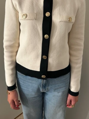 Vit kofta med svarta detaljer - Stilren vit kofta med svarta ribbade muddar och knappslå. Guldknappar framtill och två dekorativa bröstfickor. Långärmad och i mjukt stickat material som passar perfekt till jeans eller kjol.