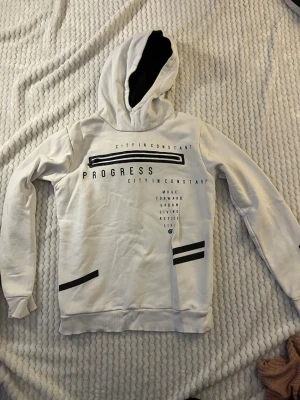 Vit hoodie med svart text och tryck - Säljer en vit hoodie med svart text och grafiska tryck på framsidan. Texten inkluderar ord som 'PROGRESS' och 'CITY IN CONSTANT'. Hoodien har en stor huva med svart innerfoder och ribbade muddar vid ärmar och nederkant. Perfekt för en avslappnad streetwear-look.