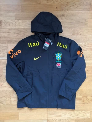 Svart Brasil Nike vindjacka med huva - Snygg svart vindjacka från Nike med Brasil-logga och sponsormärken i gult och orange. Jackan har huva, dragkedja framtill och fickor på sidorna. Perfekt för dig som gillar sportig stil och vill sticka ut med landslagskänsla.