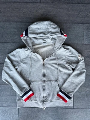Grå moncler zip up  - Snygg grå hoodie med dragkedja och huva. Ärmsluten och huvan har röd, vit och marinblå randig detalj. Två fickor framtill och dragsko i huvan. Mjuk bomullskänsla och patch på vänster ärm.