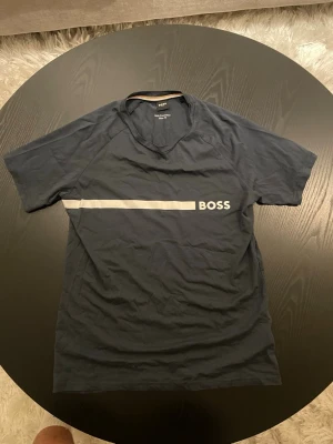 Mörkblå BOSS t-shirt - Snygg mörkblå t-shirt från BOSS med en vit horisontell linje och logga framtill. Klassisk passform och rund halsringning, perfekt för en stilren och enkel look. Storlek S