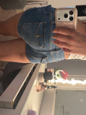 Blå denimshorts med fickor - Säljer ett par klassiska blå denimshorts med fickor både fram och bak. Shortsen har låg midja, knapp- och dragkedjestängning samt en tight passform. Perfekta för varma sommardagar och enkla att matcha med olika toppar.