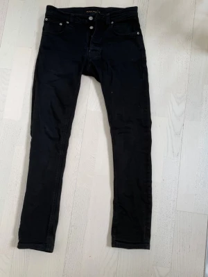 Svarta nudie jeans - Snygga svarta skinny jeans med klassisk femficksdesign och smal passform. Jeansen har bälteshällor, knappgylf och diskreta sömmar. Perfekta för en stilren och trendig look. Storlek w29 L30 och är lite rasiga på en av öglorna man sätter bälte i