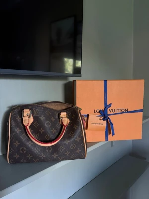 Louis Vuitton Speedy Monogram väska - Säljer en klassisk Louis Vuitton Speedy handväska i brunt monogramcanvas med beigea läderhandtag och detaljer. Väskan har guldfärgade metalldetaljer och dragkedja upptill. Ikonisk design som är lätt att matcha till olika outfits. Speedy 25!