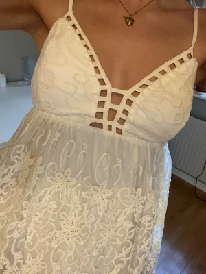Vit spetslinne med broderade detaljer - Linne i vitt med tunna axelband och djup v-ringning med dekorativa band framtill. Toppen har broderade blommor och spetsdetaljer över hela tyget samt en flowy passform. Perfekt för en somrig och fräsch look.