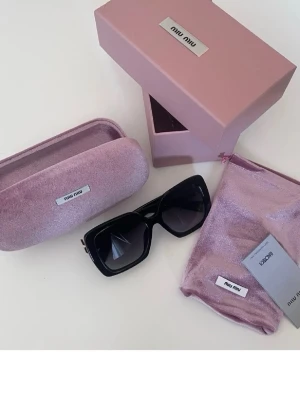 Svarta solglasögon från Miu Miu - Stora, fyrkantiga svarta solglasögon från Miu Miu med mörka glas. Kommer med ett lyxigt rosa fodral, matchande tygpåse och originalkartong. Perfekta för dig som vill ha en statement-accessoar med exklusiv känsla.